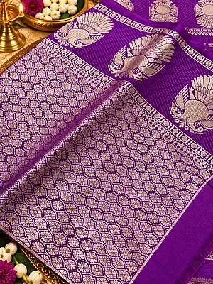 ? ROYAL PURPLE SILK SAREE A stunning royal purple silk saree featuring intricate gold zari weaving, elegant peacock motifs, and a beautifully detailed geometric floral pattern.